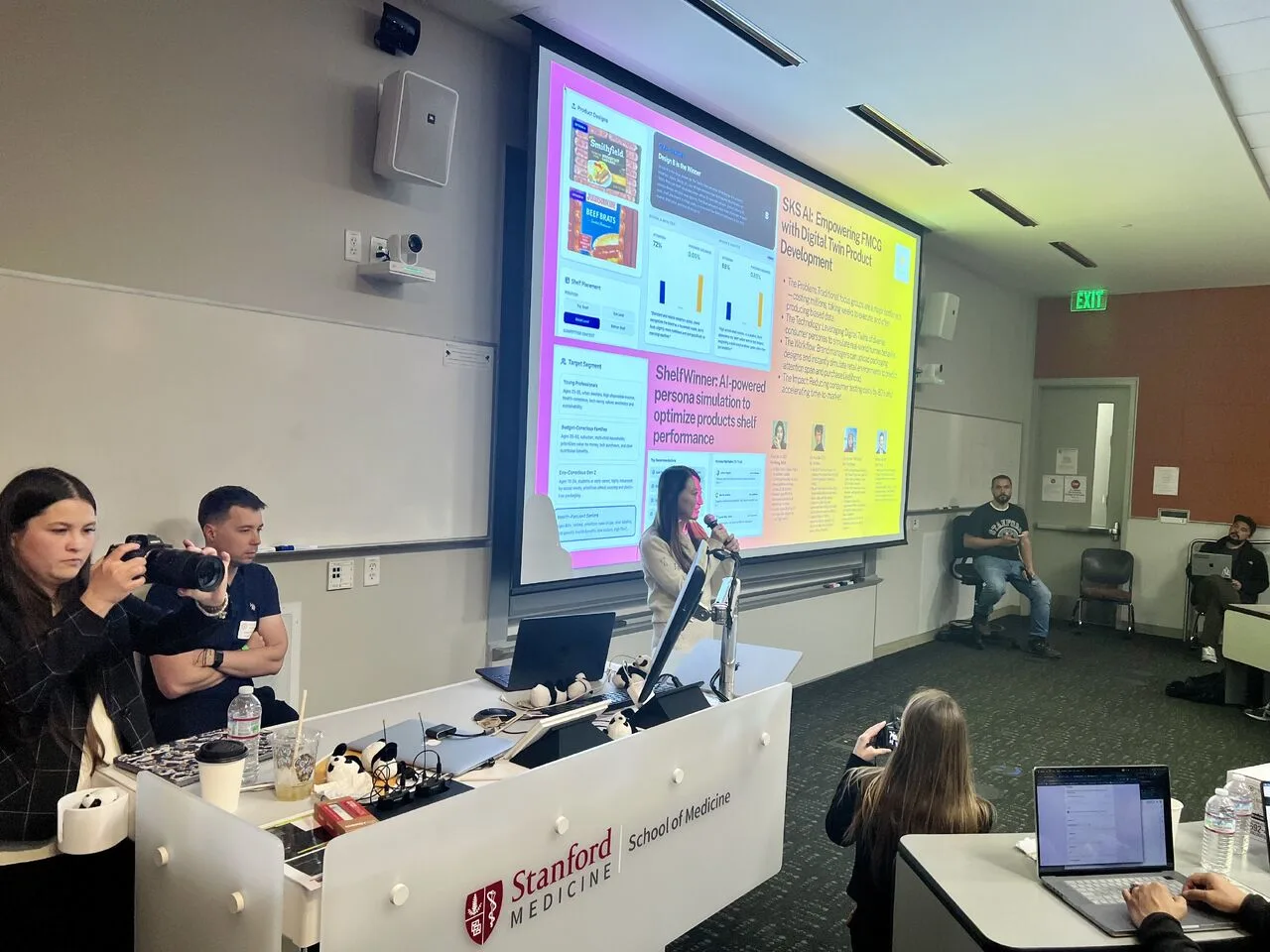 Ke Wang on stage at the Stanford × DeepMind Hackathon at Stanford School of Medicine, presenting the SKS AI SmartCart pitch with the product slide projected behind her